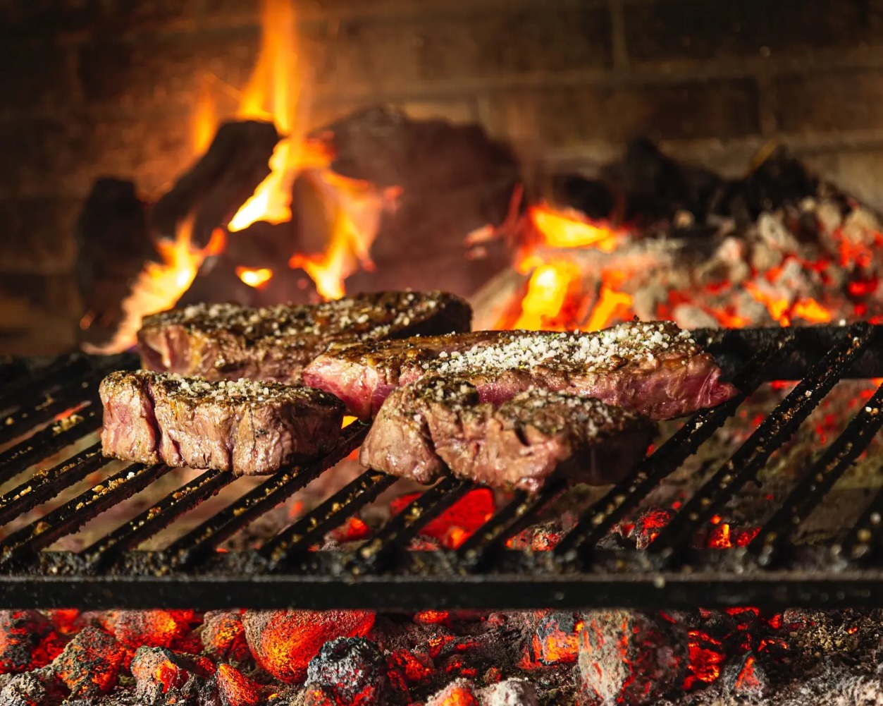 Cortes de carne cocinándose en una parrilla de carbón con fuego.
