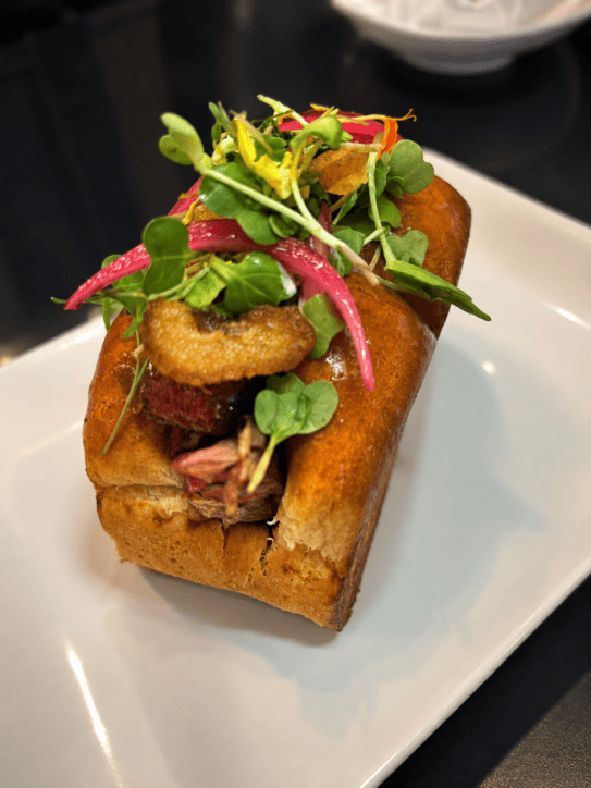 an brioche tostado y relleno de costilla desmechada con aros de cebolla y brotes.